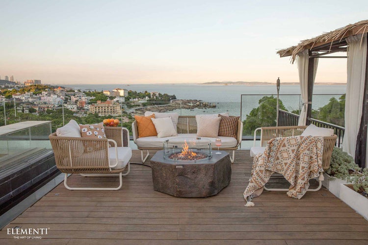Moderne Outdoor-Lounge auf Holzterrasse mit Meerblick, Seilgeflecht-Möbeln, Kissen und Gaskamin in Steinoptik mit Glasschutz. Elementi Logo.
