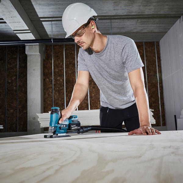 Ein Mann mit Schutzhelm und Schutzbrille arbeitet mit einer Stichsäge auf einer Holzplatte.