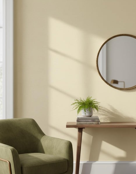 Wohnraum mit einer Wand in sanftem Gelbton, einem Sessel, einem Holztisch mit Pflanzen und einem runden Wandspiegel in einem modernen Ambiente.