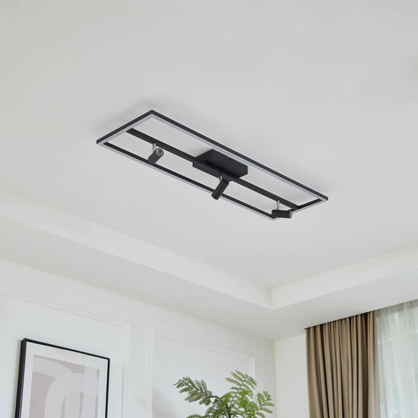 Moderne rechteckige Metall-Deckenleuchte mit drei Spots an einer Decke in einem hellen, minimalistischen Zimmer mit Pflanze und Vorhang.