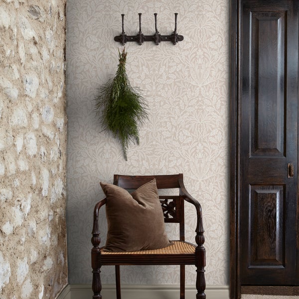 Vliestapete mit Eichenblatt- und Eicheldesign in einem hellen, rustikalen Raum mit Natursteinwand, Holzstuhl und dunkler Holztür.