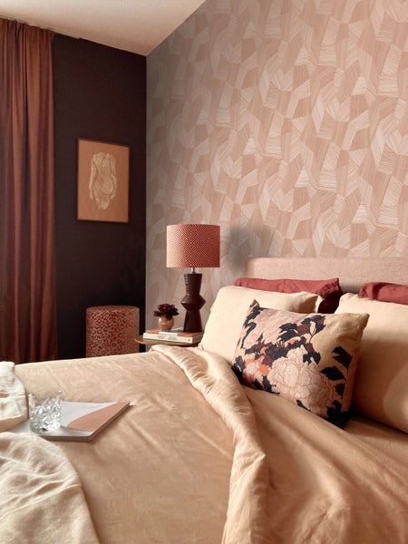 Modernes Schlafzimmer mit geometrisch gemusterter Tapete, Bett mit floralen Kissen, Nachttischlampe und dunkler Wand mit gerahmtem Bild.