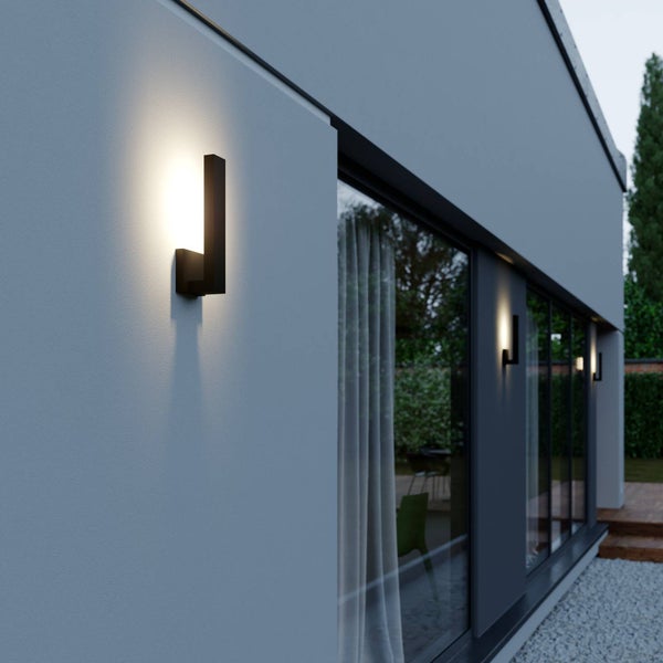 Moderne schwarze Außenwandleuchten an einer grauen Hausfassade neben großen Fensterfronten bei Dämmerung.