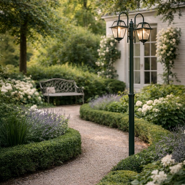 Zweiflammige nostalgische Gartenlaterne in einem gepflegten Garten mit Blumenbeeten, Hecken und einem Kiesweg vor einem Wohnhaus.