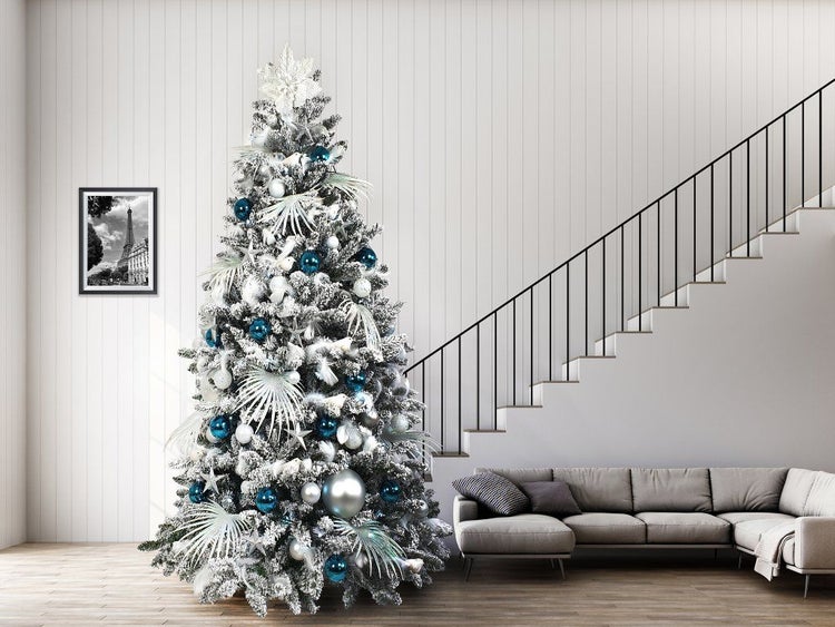 Ein geschmückter Weihnachtsbaum mit Schneeoptik, verziert mit Kugeln und Sternen, steht in einem modern eingerichteten Wohnzimmer mit Treppe.