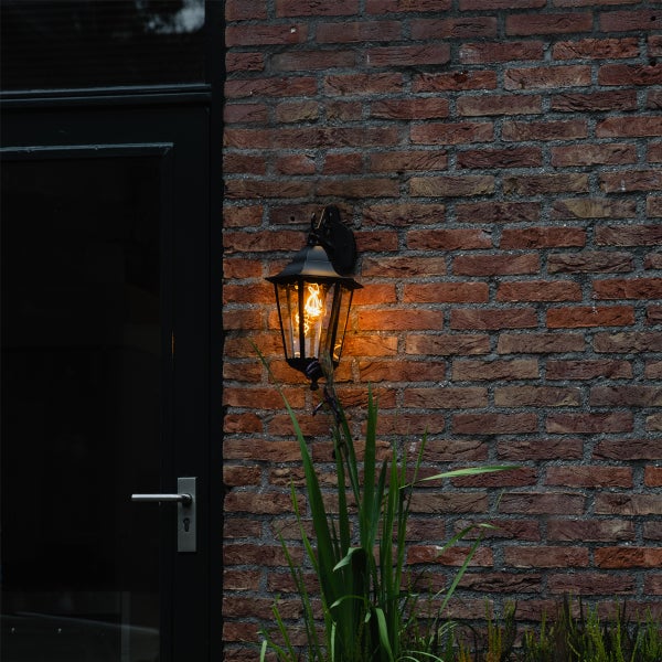 Klassische schwarze Außenwandleuchte mit warmem Licht, montiert an einer Backsteinwand neben einer Tür.