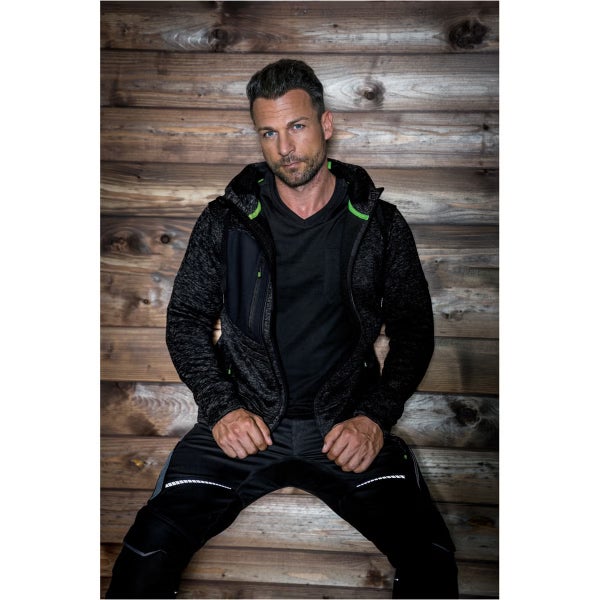 Mann in dunkler Strickfleecejacke mit Kapuze und neongrünen Akzenten sowie schwarzer Arbeitshose vor einer Holzwand.