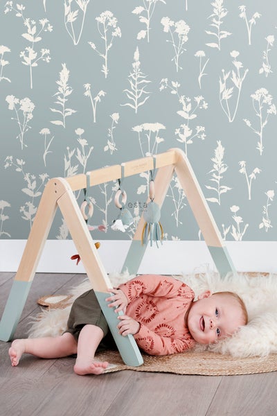 Baby liegt unter einem Spielbogen mit Spielzeug auf einem Schaffellteppich vor einer Wand mit Pflanzenmotiven