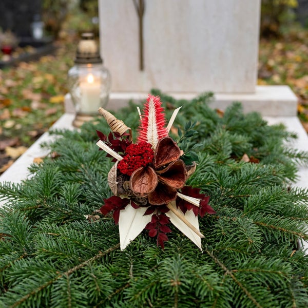 Grabschmuck aus Tannengrün mit Trockenblumen und einer Grabkerze auf einem hellen Grabstein.