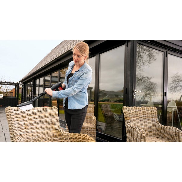 Eine Frau reinigt einen Gartenstuhl aus Korbgeflecht mit einer Waschbürste für Hochdruckreiniger auf einer Terrasse vor einem Wintergarten.
