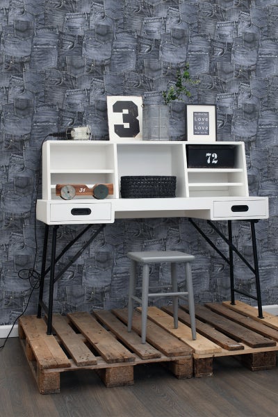 Schreibtisch mit Regalen und Hocker auf Holzpalette vor einer Wand mit Jeansmuster