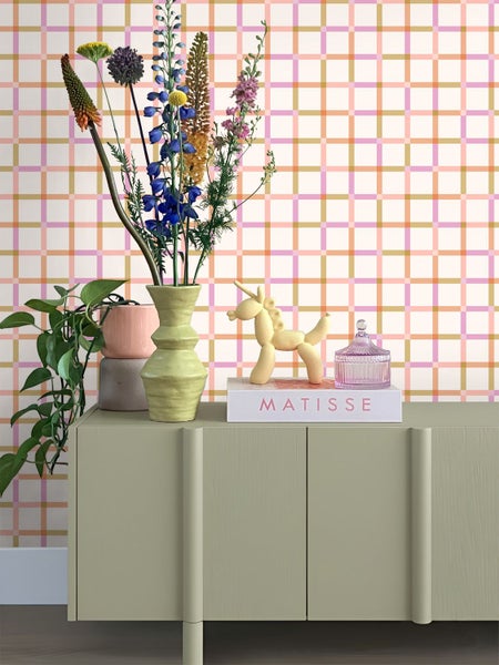 Karierte Tapete mit Linien in Rosa, Orange und Gold hinter einem grünen Sideboard mit Blumen und Deko-Objekten.