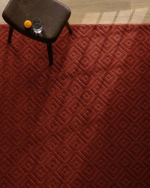 Roter Teppich mit geometrischem Rautenmuster, darauf ein dunkler Stuhl mit einem Glas Wasser und einer Orange auf hellem Holzboden.