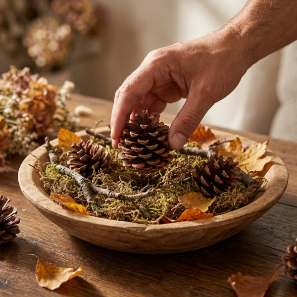 Eine Hand platziert einen Kiefernzapfen in eine dekorative Holzschale gefüllt mit Moos, Zweigen und Herbstblättern auf einem Holztisch.