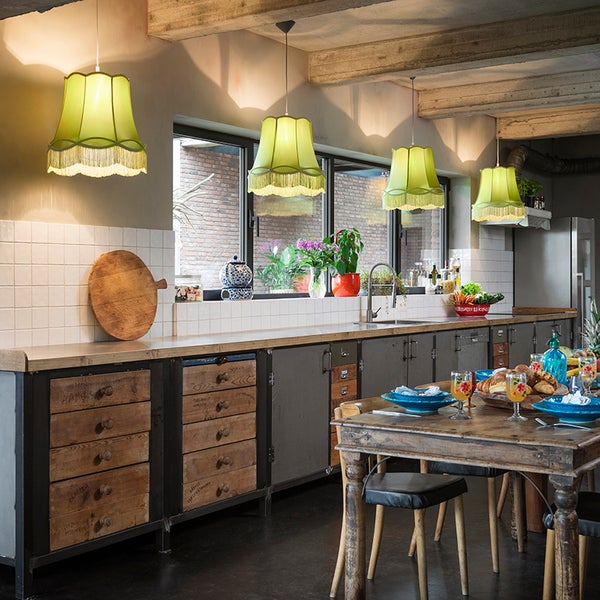 Vier grüne Pendelleuchten mit Fransen über einer langen Küchentheke im Industriestil und einem Holzesstisch in einem rustikalen Raum.