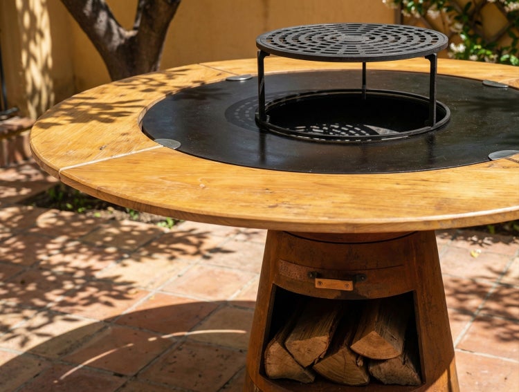 Runder Grilltisch mit Holztischplatte, integrierter Feuerstelle, Grillring und Holzlagerfach im Sockel auf einer Terrasse.