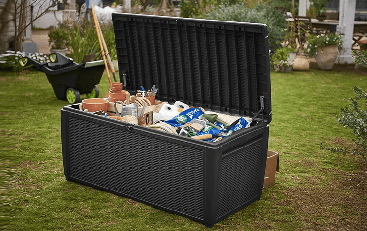 Offene schwarze Gartenbox in Rattan-Optik auf einer Rasenfläche, gefüllt mit Gartenwerkzeugen, Blumentöpfen und Säcken mit Erde.
