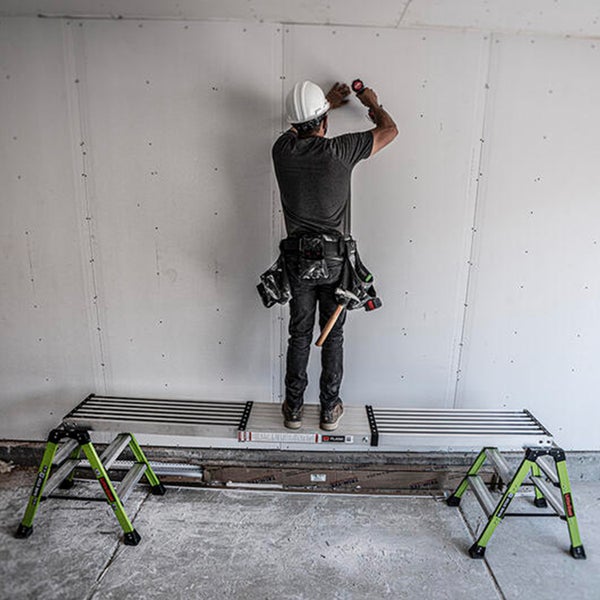 Handwerker mit Schutzhelm steht auf einer robusten Aluminium Arbeitsplattform zwischen zwei grünen Arbeitsböcken bei der Wandmontage.