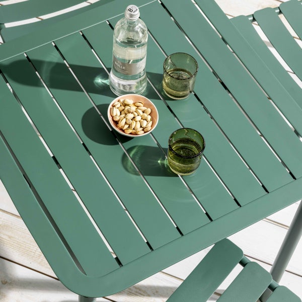 Grüner Metall-Gartentisch mit Lattendesign, Wasserflasche, zwei Gläsern und Pistazienschale im Sonnenlicht auf einer Holzterrasse.