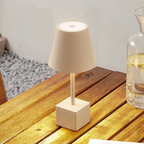Kabellose LED-Tischleuchte in Beige mit konischem Schirm und quadratischem Standfuß auf einem Holztisch im Außenbereich.