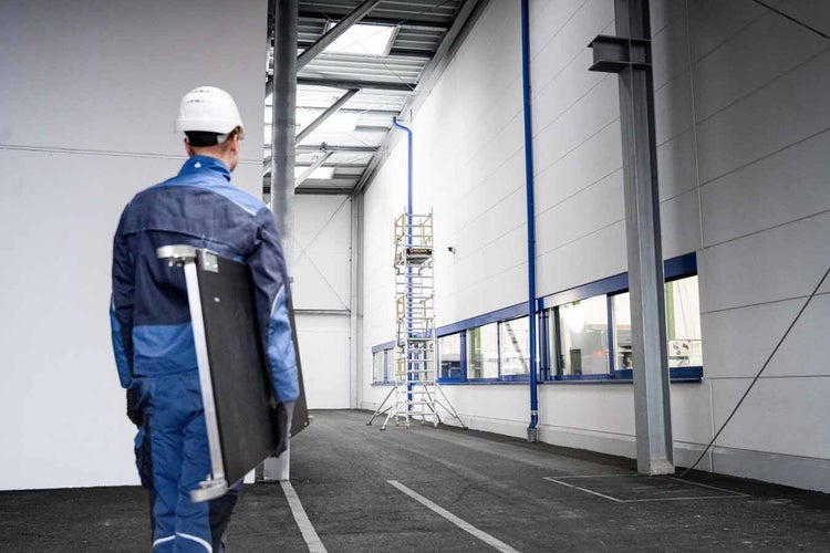 Ein Arbeiter in Arbeitskleidung trägt eine Platte durch eine helle Industriehalle. Im Hintergrund steht ein hohes fahrbares Gerüst.