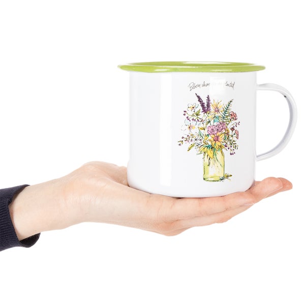 Emaille-Tasse mit Blumenmotiv in einem Glas, Aufschrift Bloom where you are planted und grünem Rand, auf einer Hand präsentiert.