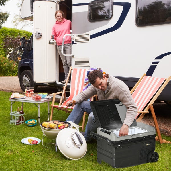 Tragbare elektrische Kompressor-Kühlbox mit Rollen in einer organisierten Camping-Szene vor einem Wohnmobil mit Grill und Liegestühlen.