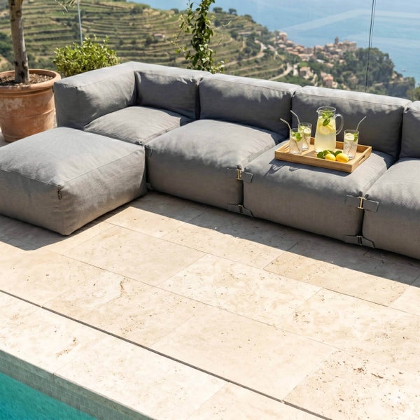 Modulare Gartenlounge aus strukturiertem Stoff auf einer Terrasse mit Natursteinfliesen und Blick auf eine Küstenlandschaft.