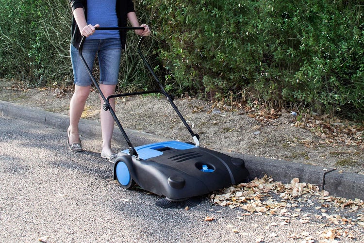 Eine Person bedient eine handgeführte Kehrmaschine auf Asphalt, um Laub entlang einer Bordsteinkante aufzukehren.