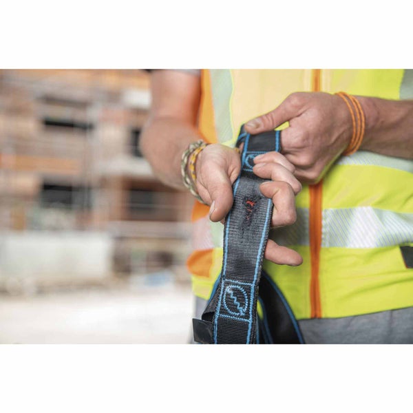 Person in Warnweste prüft einen schwarzen Sicherheitsgurt mit MAS Logo und blauen Nähten auf einer Baustelle.