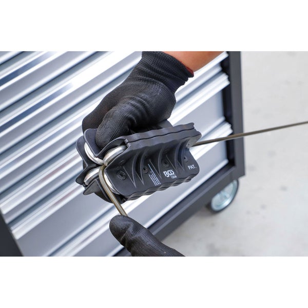 Ein BGS technic Drahtrichtwerkzeug wird von Händen in Handschuhen genutzt, um einen Metalldraht vor einem Werkzeugwagen aus Metall zu richten.