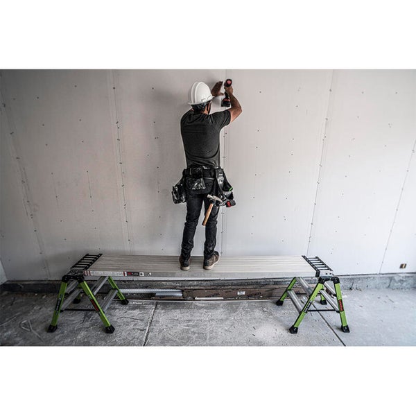 Handwerker mit Schutzhelm steht auf einer robusten Arbeitsplattform mit Gerüstböcken und montiert Gipskartonplatten an einer Wand.