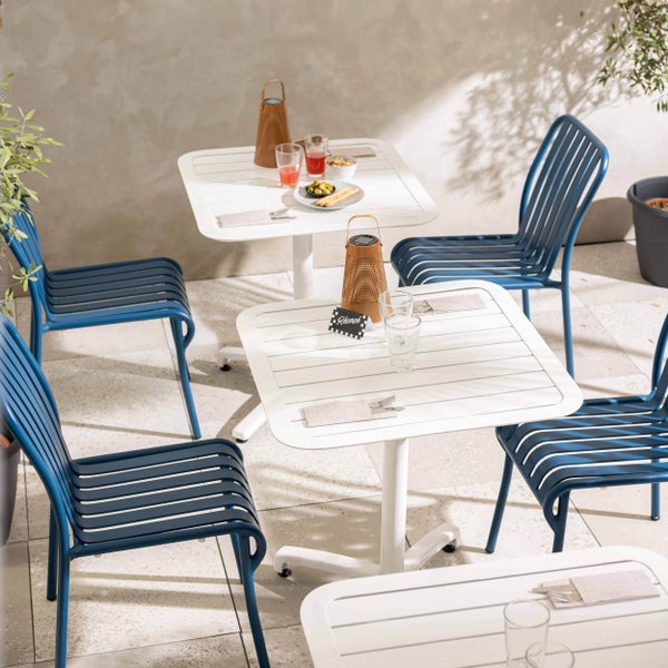 Blauer Bistrostuhl aus Metall mit Lattendesign auf einer Terrasse mit weißen Tischen und hellen Steinplatten.