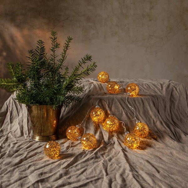 Dekorative Szene mit einer Lichterkette in Kugelform und einer Grünpflanze in einem goldenen Topf auf einem Stofftisch