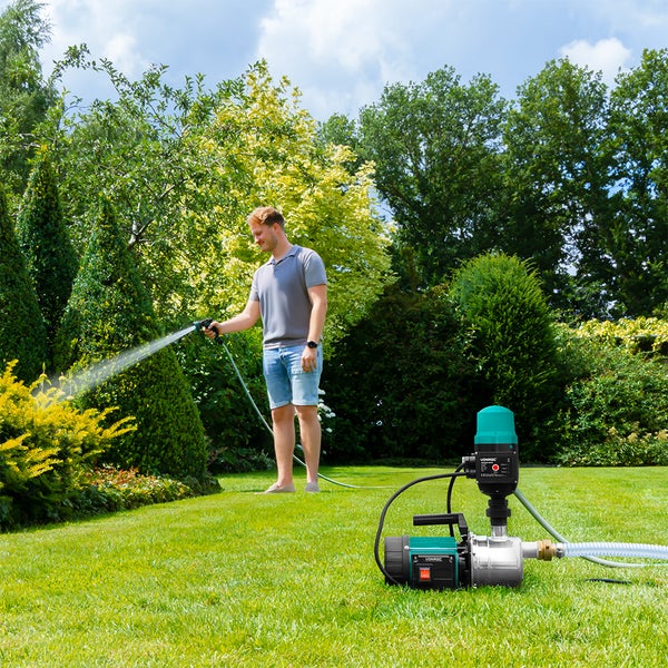 Ein Mann bewässert Sträucher in einem Garten mit einer Spritzpistole, während im Vordergrund ein Hauswasserautomat auf dem Rasen steht.