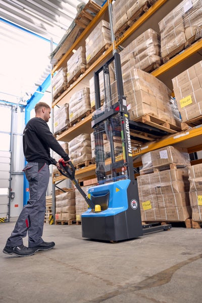 Mitarbeiter bedient einen blauen HanseLifter Elektro-Hochhubwagen in einem Lagerhaus mit Hochregalen voller Kartons.