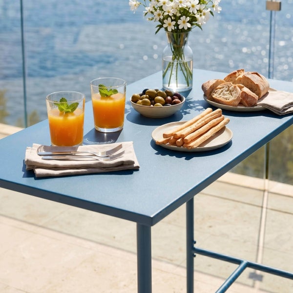 Moderner blauer Bartisch aus Metall im Freien mit Gläsern, Oliven, Brot und Blumen auf einer hellen Terrasse mit Meerblick.