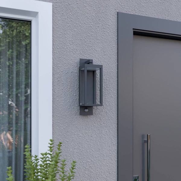 Moderne rechteckige Außenwandleuchte mit Bewegungsmelder montiert an einer Hausfassade neben einer Tür und einem Fenster.