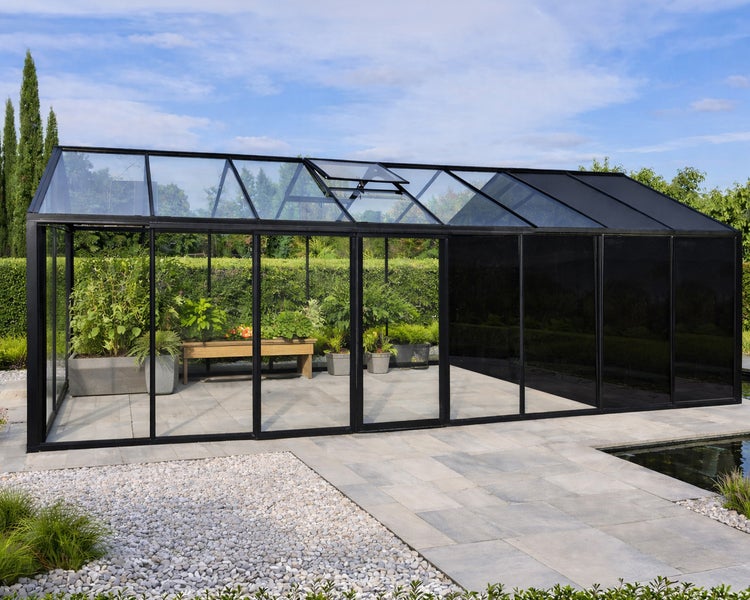 Modernes, geräumiges Gewächshaus mit schwarzem Metallrahmen und Glaswänden, halbseitig getönt, auf einer Steinterrasse mit Pflanzen und Teich.