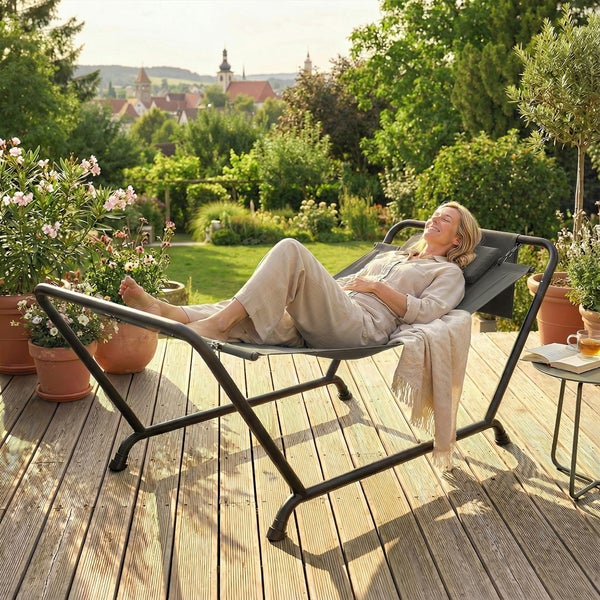 Eine Frau entspannt in einer grauen Gartenliege auf einer sonnigen Holzterrasse, umgeben von Topfpflanzen und Gartenblick.