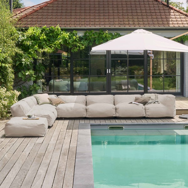 Großzügige Gartenlounge auf einer Holzterrasse am Pool, bestehend aus einem beigefarbenen Ecksofa und einem Hocker.