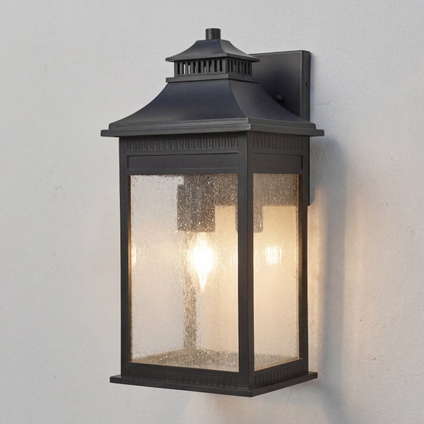 Schwarze Außenwandleuchte im Laternendesign mit klarem, strukturiertem Glas und eingeschaltetem Leuchtmittel.