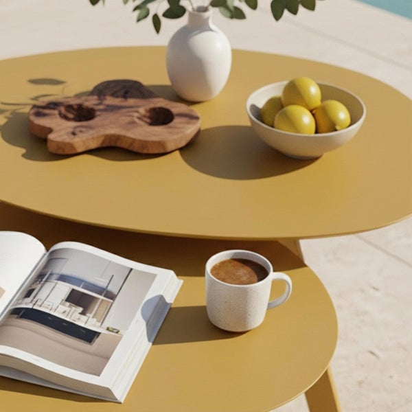 Zwei runde Beistelltische für den Außenbereich in Senfgelb, dekoriert mit Vase, Holzbrett, Zitronenschale, Buch und Kaffeetasse auf einer Terrasse.