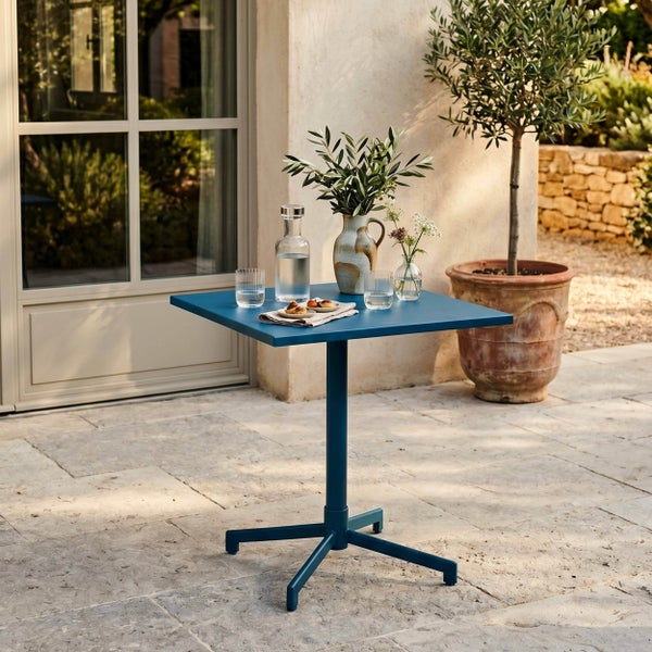 Quadratischer Bistrotisch aus Metall in Blau auf einer Terrasse mit Steinfußboden, dekoriert mit einer Wasserkaraffe und einer Blumenvase.