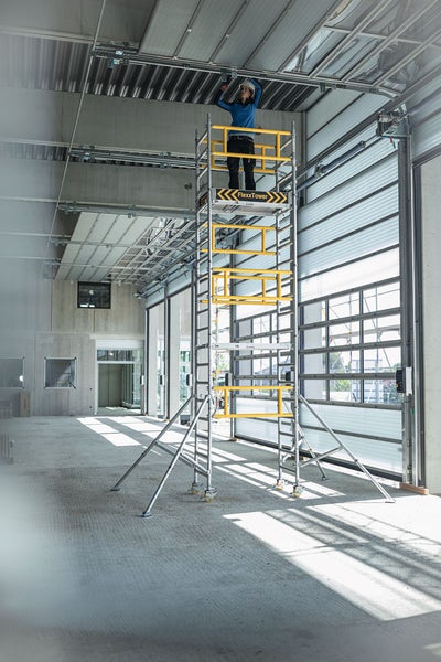 Person auf einem Rollgerüst beim Arbeiten an der Decke in einem hellen, geräumigen Raum.