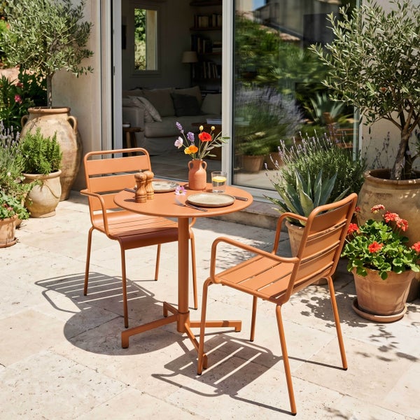 Modernes Gartenmöbel-Set mit rundem Metalltisch und zwei Stühlen in Terracotta auf einer Steinterrasse, umgeben von verschiedenen Topfpflanzen.