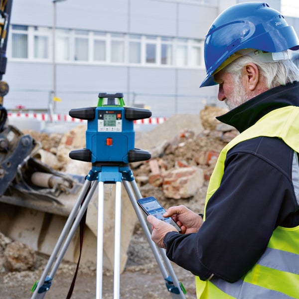 Handwerker mit Schutzhelm und Warnweste bedient Bosch Rotationslaser auf einem Stativ mit seinem Smartphone auf einer Baustelle.