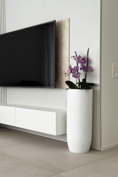 Modernes Wohnzimmer mit wandmontiertem Fernseher, schwebendem weißem Lowboard und hohem weißem Pflanzgefäß mit lila Orchidee auf Fliesenboden.