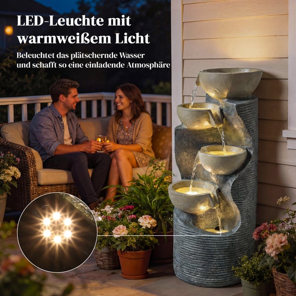Beleuchteter Gartenbrunnen in Steinoptik mit warmweißem Licht auf einer Terrasse mit sitzendem Paar im Hintergrund in Abendstimmung.