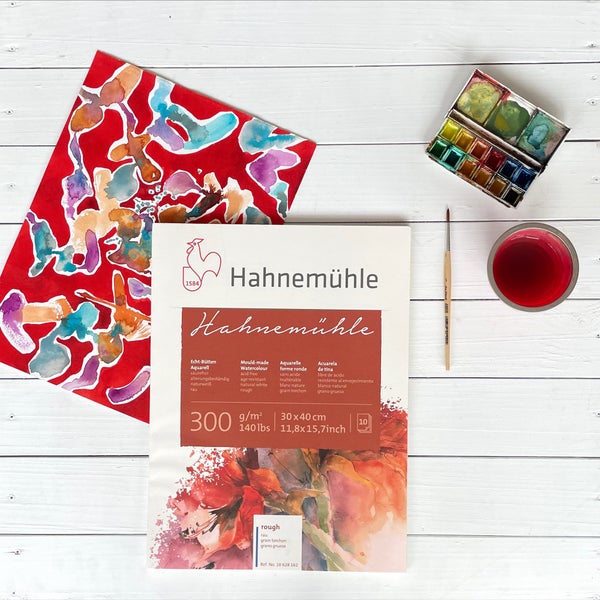 Hahnemühle Aquarellblock 300 Gramm pro Quadratmeter, 30 mal 40 Zentimeter, 10 Blatt, raue Oberfläche, mit Aquarellfarben und Pinsel auf Holztisch.